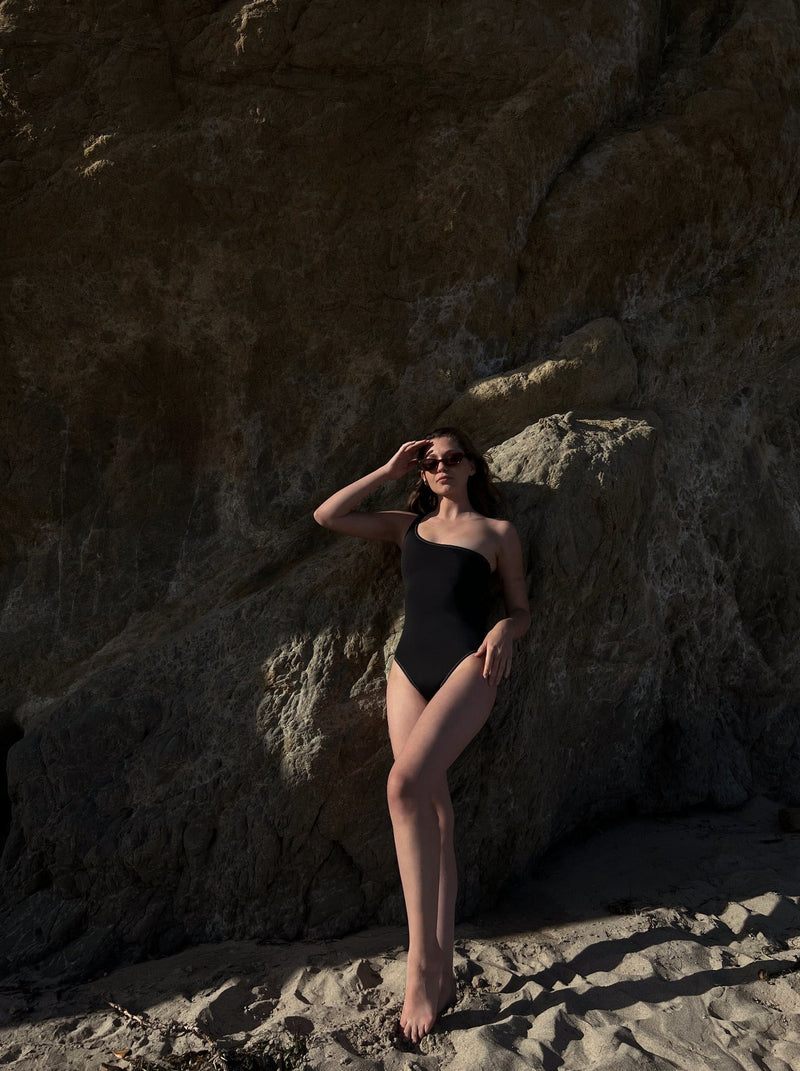 woman leaning on a boulder in black one shoulder one pice swimsuit and sunglasses